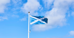 The Scottish flag blows in the wind against a blue sky, with light white clouds