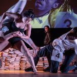 A culturally diverse group of dancers performs among piles of books. They wear costumes with large numbers pinned to them, suggesting an audition or competition. Their movements are dynamic and physical, with one dancer holding another’s leg as others lean and bend in varied, expressive poses.