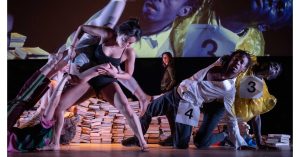 A culturally diverse group of dancers performs among piles of books. They wear costumes with large numbers pinned to them, suggesting an audition or competition. Their movements are dynamic and physical, with one dancer holding another’s leg as others lean and bend in varied, expressive poses.