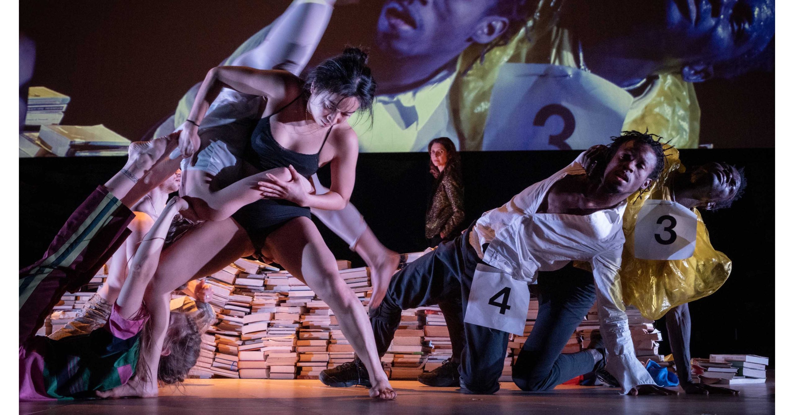 A culturally diverse group of dancers performs among piles of books. They wear costumes with large numbers pinned to them, suggesting an audition or competition. Their movements are dynamic and physical, with one dancer holding another’s leg as others lean and bend in varied, expressive poses.