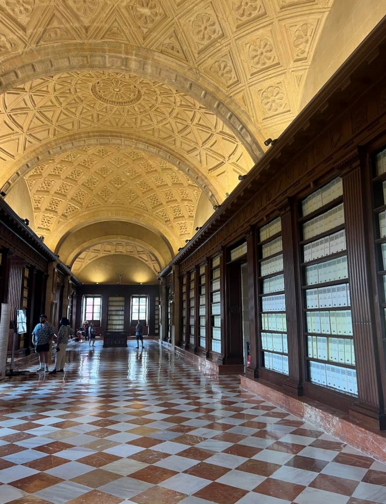 Long interior hall of the Archivo General de Indias with a richly decorated vaulted ceiling, checkerboard marble floor, tall wooden shelving filled with archival volumes, and a few visitors walking through the space.