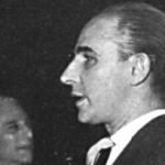 Profile half-length photograph of a man facing to the left wearing a dark jacket and tie, holding a cigarette in his hand, with a white handkerchief in his jacket breast pocket. The man has a prominent straight nose, slicked back hair and his tie forms a slight arch from the knot. In the background, on the left side of the image, there is another half-length frontal portrait of a man. He has white hair, also wears a jacket and tie, is looking to the right, and is smiling.