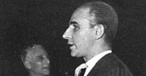 Profile half-length photograph of a man facing to the left wearing a dark jacket and tie, holding a cigarette in his hand, with a white handkerchief in his jacket breast pocket. The man has a prominent straight nose, slicked back hair and his tie forms a slight arch from the knot. In the background, on the left side of the image, there is another half-length frontal portrait of a man. He has white hair, also wears a jacket and tie, is looking to the right, and is smiling.
