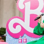 Two women sit on a small outdoor stage holding microphones and speaking to each other. One wears a bright pink dress and smiles while listening; the other wears a green dress and speaks into the microphone. Behind them is a large pink decorative sign, and two water bottles sit on a table between them. The setting appears to be a public talk or interview event.