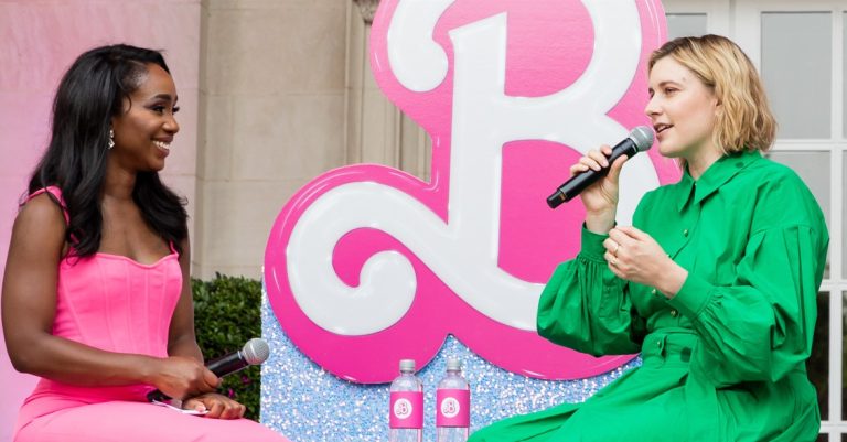 Two women sit on a small outdoor stage holding microphones and speaking to each other. One wears a bright pink dress and smiles while listening; the other wears a green dress and speaks into the microphone. Behind them is a large pink decorative sign, and two water bottles sit on a table between them. The setting appears to be a public talk or interview event.