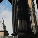 Stone statue viewed from behind beneath tall Gothic arches and dark stone columns, framing a bright blue sky with clouds
