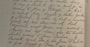Close-up of a historical handwritten manuscript page in cursive ink, showing dense text on aged paper, held open by a person’s hand at the bottom edge.