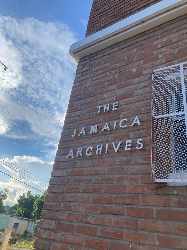 Exterior brick wall of The Jamaica Archives with white lettering reading ‘The Jamaica Archives,’ a barred window to the right, and blue sky with clouds in the background.