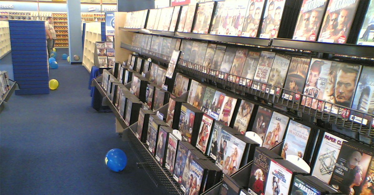 Interior of a video rental store aisle with rows of DVD cases displayed on wall-mounted racks. Blue carpeted floor with several loose balloons scattered along the aisle. Shelving and displays extend into the background, with one person partially visible near the back of the store.