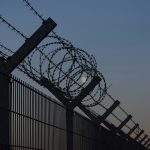 Barbed wire and razor wire coiled along the top of a tall metal security fence silhouetted against a dusk sky with the moon visible in the background.