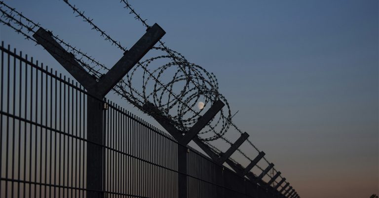 Barbed wire and razor wire coiled along the top of a tall metal security fence silhouetted against a dusk sky with the moon visible in the background.