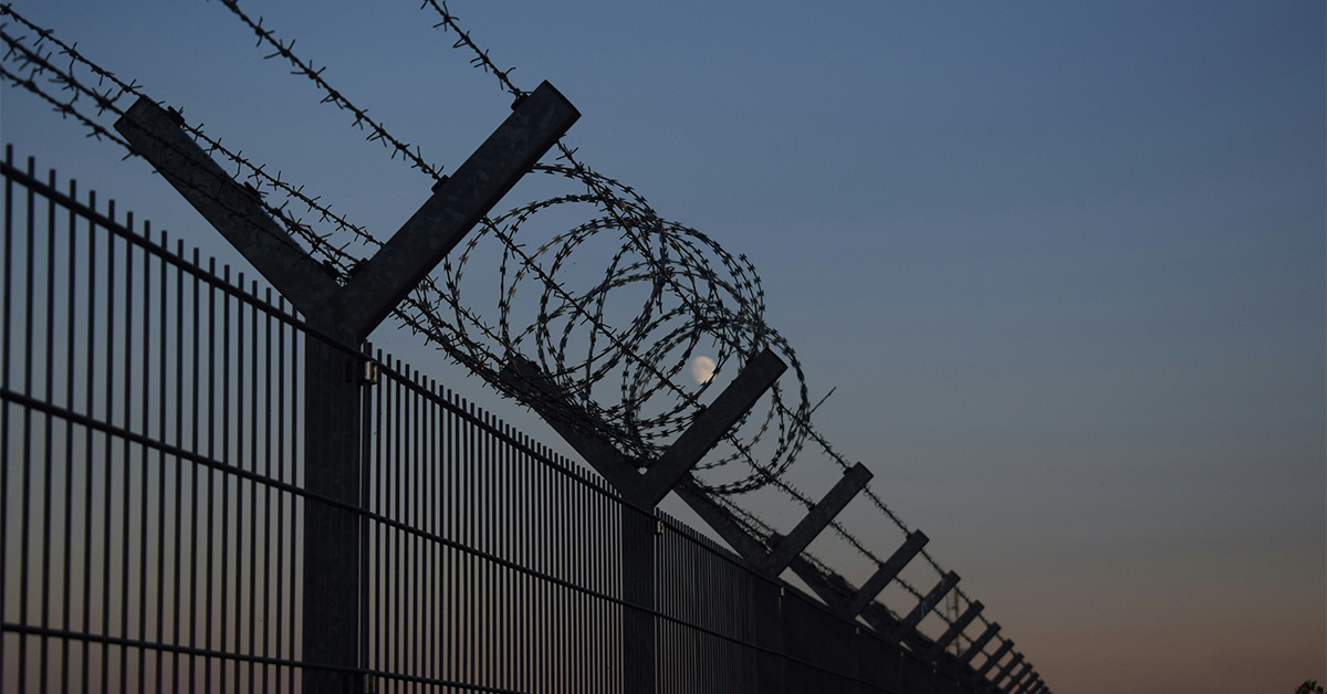 Barbed wire and razor wire coiled along the top of a tall metal security fence silhouetted against a dusk sky with the moon visible in the background.