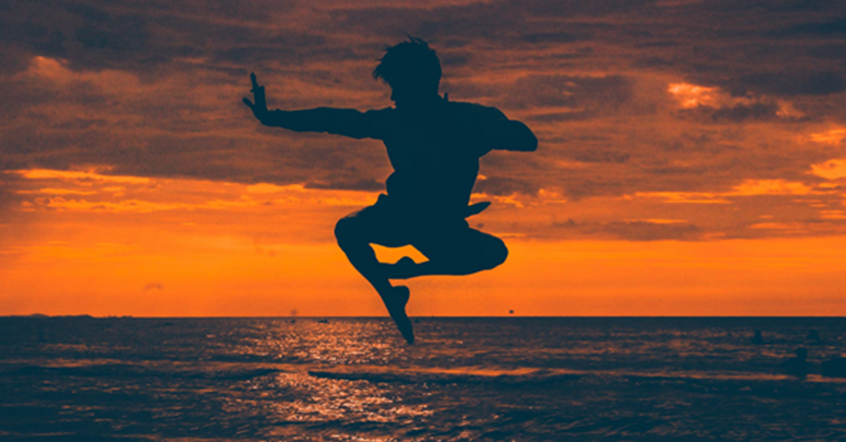 Silhouetted martial artist mid-air against a sunset sky over the sea, holding a poised, balanced stance that emphasises calm, control, and intention rather than force.
