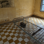 The image shows a sparse, worn room that appears to be a former prison cell. The walls are yellowed and heavily stained, with patches of peeling paint and dark discoloration near the bottom. The floor has a checkerboard pattern of tan and white tiles. In the center of the room is a simple metal bed frame with a grid base and no mattress. On top of the bed frame sits a small metal box. Attached to the frame are metal shackles, suggesting restraints were used. The room has a barred window on the right side, allowing some daylight to enter, casting shadows on the floor. The overall atmosphere feels stark, somber, and austere.
