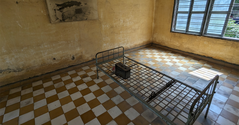 The image shows a sparse, worn room that appears to be a former prison cell. The walls are yellowed and heavily stained, with patches of peeling paint and dark discoloration near the bottom. The floor has a checkerboard pattern of tan and white tiles. In the center of the room is a simple metal bed frame with a grid base and no mattress. On top of the bed frame sits a small metal box. Attached to the frame are metal shackles, suggesting restraints were used. The room has a barred window on the right side, allowing some daylight to enter, casting shadows on the floor. The overall atmosphere feels stark, somber, and austere.