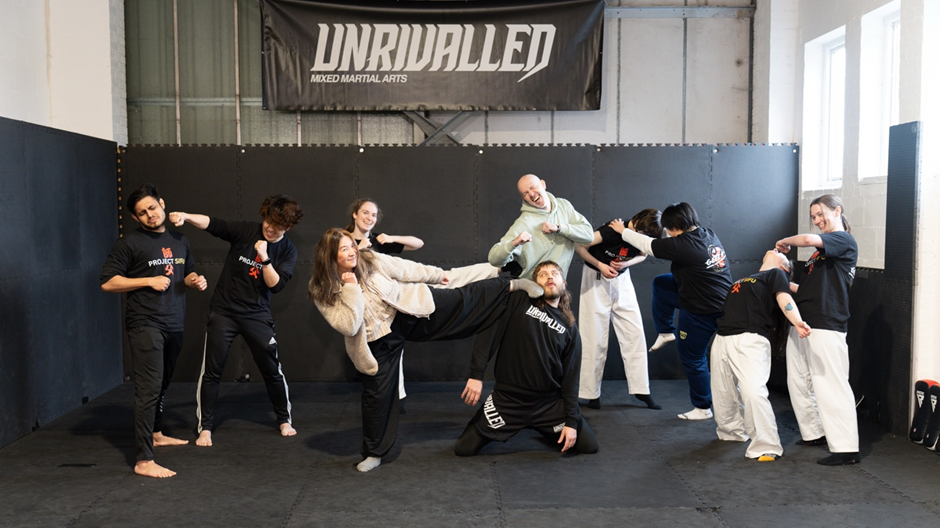 A mixed group of young people and adults in a martial arts gym collaboratively re-enact a choreographed fight scene, using playful, exaggerated movements that emphasise participation, balance, and shared creativity rather than realism.
