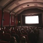 A dimly lit movie theater filled with seated audience members facing a large, brightly glowing screen. Rows of red upholstered seats extend toward the front, and the ceiling features curved architectural details with recessed panels. The atmosphere suggests a film screening in progress.