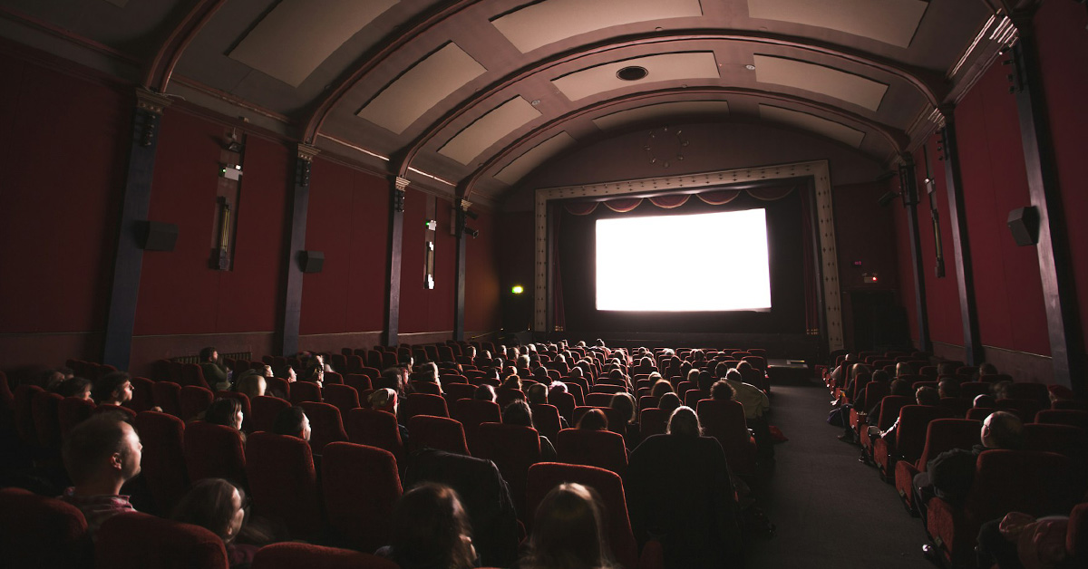 A dimly lit movie theater filled with seated audience members facing a large, brightly glowing screen. Rows of red upholstered seats extend toward the front, and the ceiling features curved architectural details with recessed panels. The atmosphere suggests a film screening in progress.