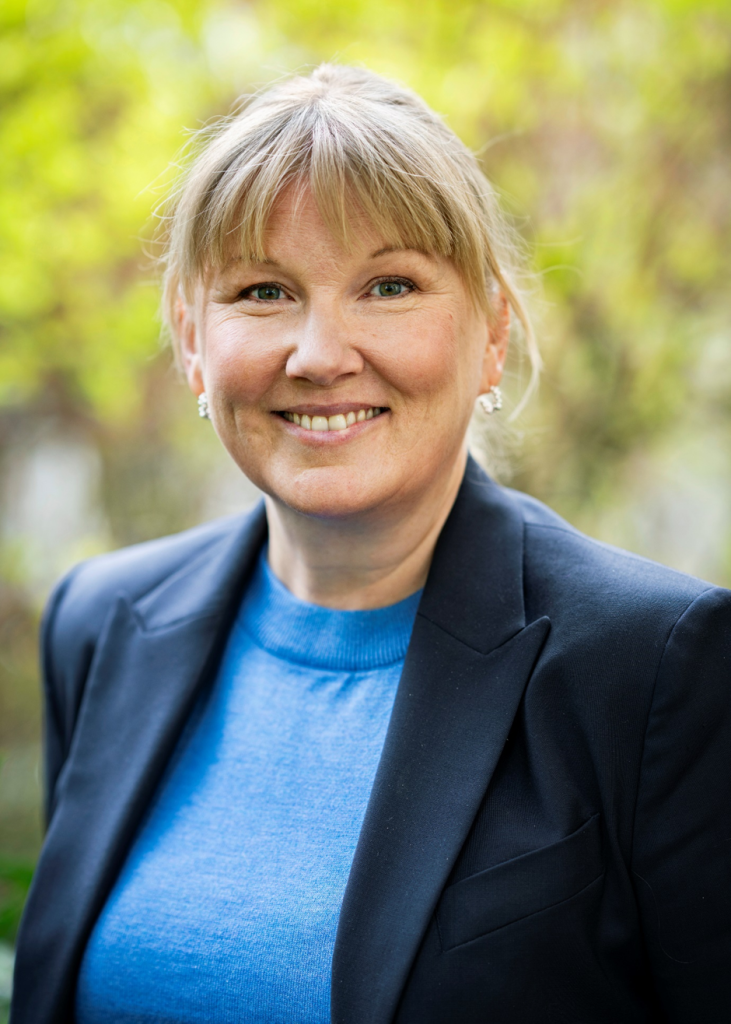 A woman with blonde hair, tied behind her head, smiles at the camera. She is wearing a black blazer and a blue jumper.