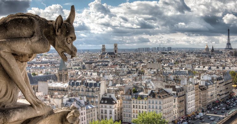 Stone gargoyle perched on Notre-Dame Cathedral overlooking the dense rooftops of Paris, with the Seine River running alongside the city and the Eiffel Tower visible in the distance beneath a dramatic, cloud-filled sky.
