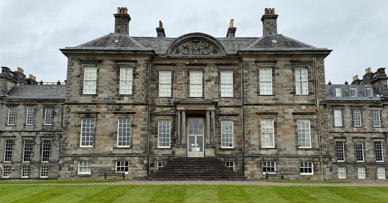 A large historic stone mansion with symmetrical architecture, tall sash windows, and a central staircase leading to the main entrance. The building has a gray weathered facade, multiple chimneys, and ornate detailing above the center section. A wide green lawn stretches across the foreground under an overcast sky.
