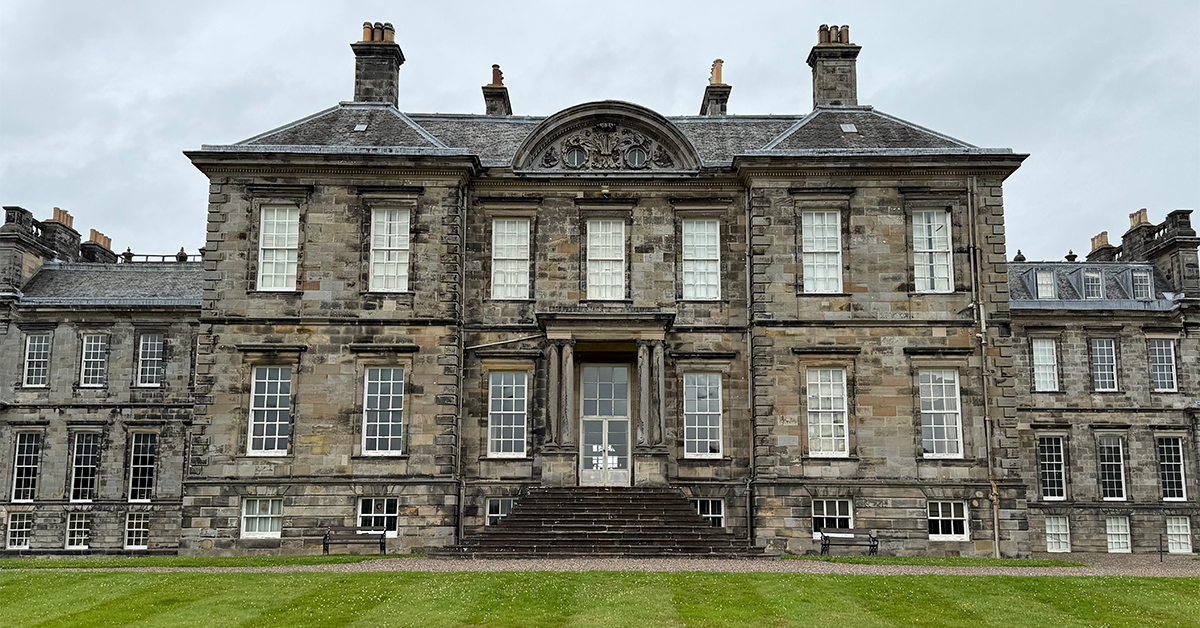 A large historic stone mansion with symmetrical architecture, tall sash windows, and a central staircase leading to the main entrance. The building has a gray weathered facade, multiple chimneys, and ornate detailing above the center section. A wide green lawn stretches across the foreground under an overcast sky.