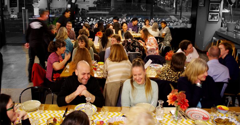 A crowded communal dinner scene inside a cozy restaurant or café. Dozens of people are seated at long shared tables, eating and chatting in a lively social atmosphere. The foreground shows smiling diners with plates, glasses, and flower arrangements on patterned table runners. The background includes large front windows looking out onto a nighttime street. The image has warm indoor lighting, with some motion blur from people walking, giving it an energetic, candid feel.