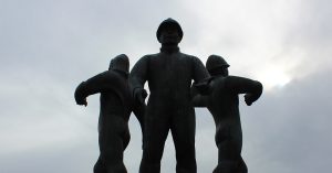 Low-angle view of a dark bronze memorial sculpture featuring three offshore oil workers standing back-to-back against a cloudy sky, silhouetted by sunlight breaking through the clouds.