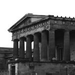 A black-and-white photo of a large neoclassical stone building with tall fluted columns, a triangular pediment, and ornate detailing along the roofline. The structure has a monumental, temple-like appearance, with dark shadows emphasizing the architectural features. Bushes and smaller adjoining sections of the building are visible in the foreground and left side.