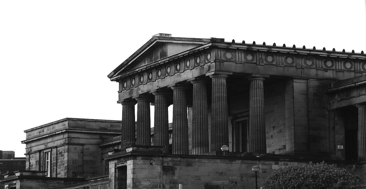 A black-and-white photo of a large neoclassical stone building with tall fluted columns, a triangular pediment, and ornate detailing along the roofline. The structure has a monumental, temple-like appearance, with dark shadows emphasizing the architectural features. Bushes and smaller adjoining sections of the building are visible in the foreground and left side.