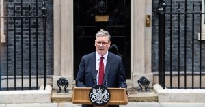 **Alt text:** A man in a dark suit and red tie stands behind a lectern with microphones, delivering a speech outside a black front door set in a brick government building. Iron railings flank the entrance, and an official crest is displayed on the front of the podium.