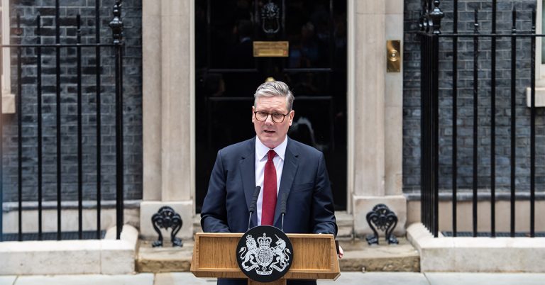 **Alt text:** A man in a dark suit and red tie stands behind a lectern with microphones, delivering a speech outside a black front door set in a brick government building. Iron railings flank the entrance, and an official crest is displayed on the front of the podium.