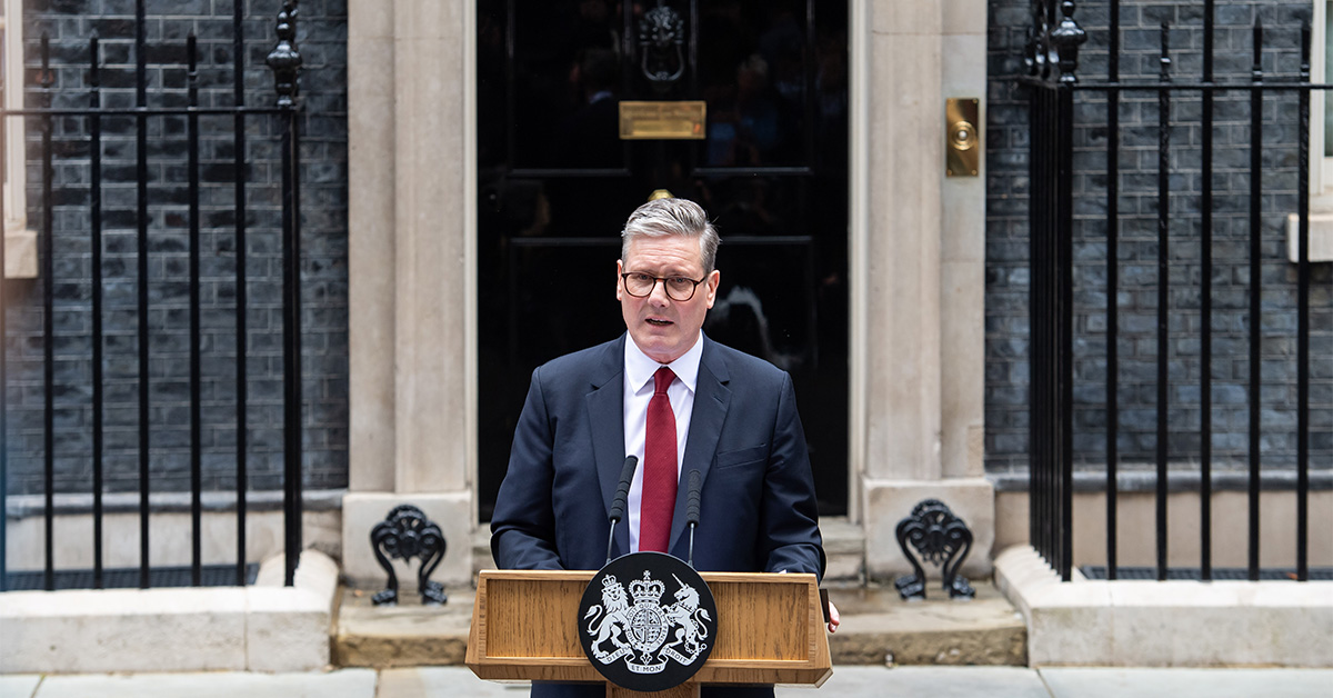 **Alt text:** A man in a dark suit and red tie stands behind a lectern with microphones, delivering a speech outside a black front door set in a brick government building. Iron railings flank the entrance, and an official crest is displayed on the front of the podium.