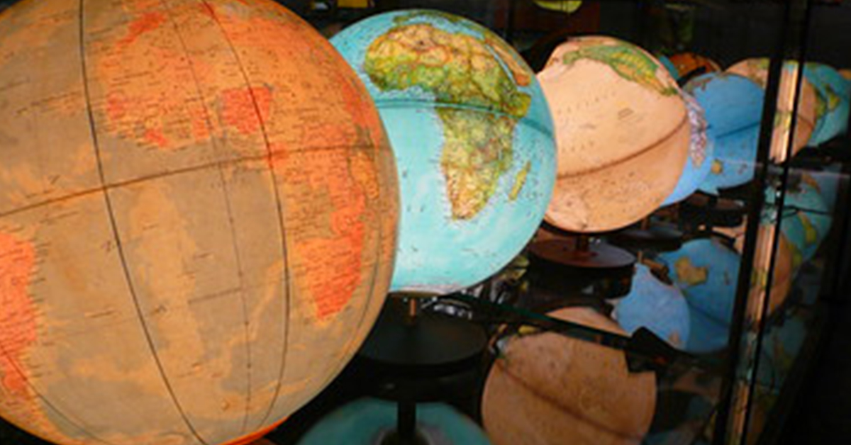 **Alt text:** A row of illuminated classroom globes displayed on stands, with a large globe in the foreground and several others receding into the background. The globes show different map styles and colors, creating a warm, softly lit scene.