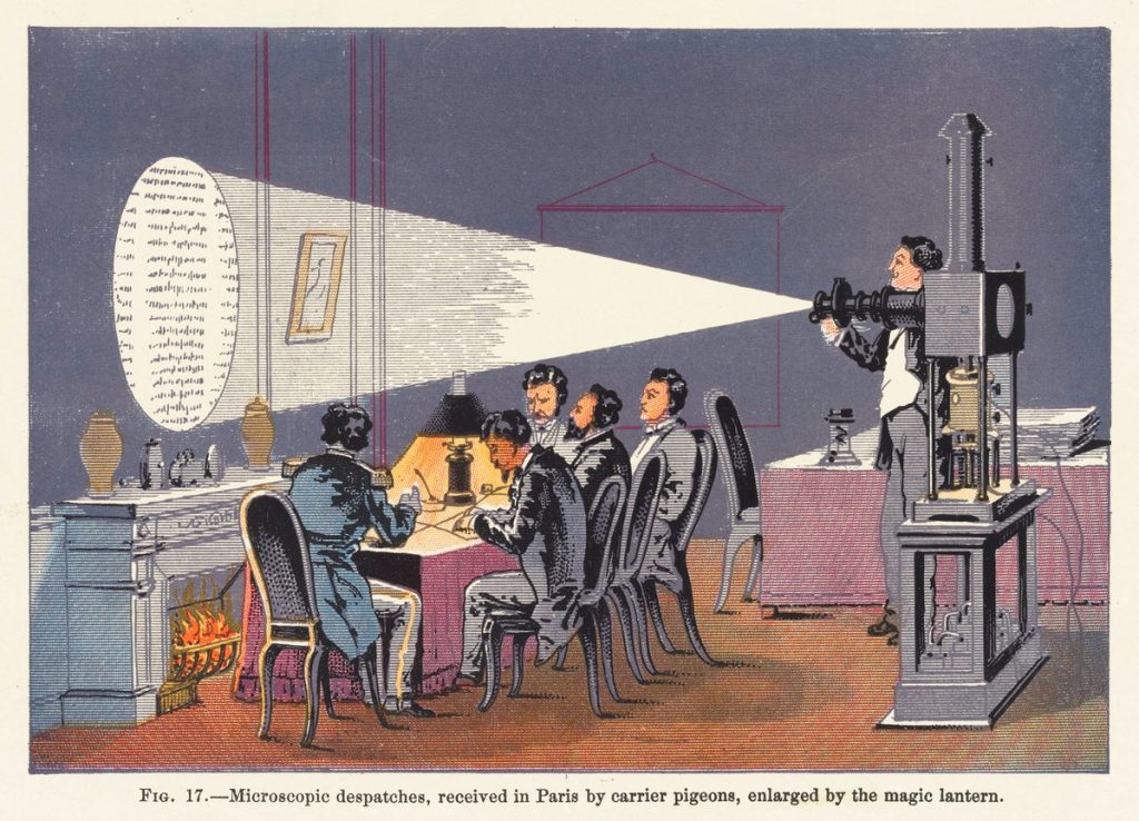 Five men in nineteenth-century tail coats are reading text projected onto a wall over a fireplace by a sixth man using a magic lantern.
