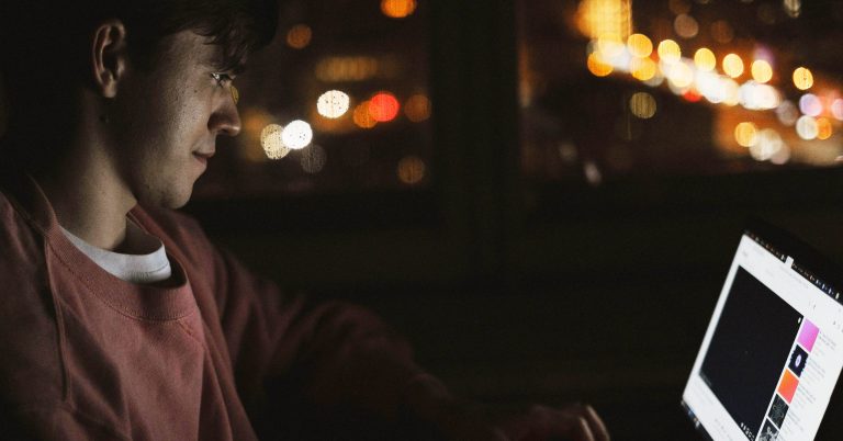 Photograph of a man in a darkly lit room on the computer. The background features distorted light from an outside building.