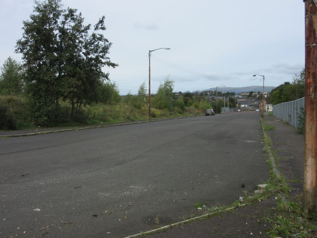 An empty wide road lined with trees, utility poles, and fencing stretches toward distant hills under an overcast sky. A single parked car is visible farther down the street.