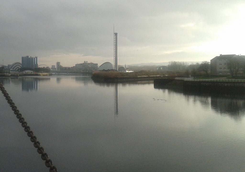A calm river or harbor reflects a cloudy sky, with modern buildings and a tall observation tower on the opposite bank. Sunlight breaks through the clouds on the right, creating bright reflections on the water.