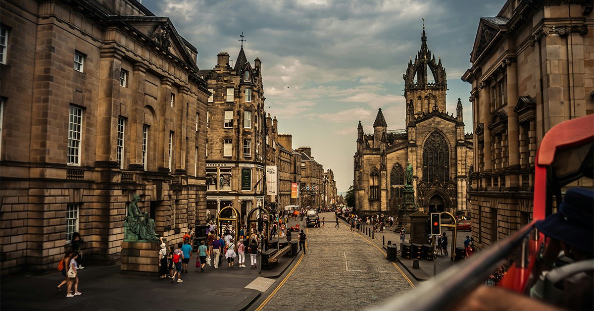 **Alt text:** A wide view down a cobblestone street in Edinburgh's historic city center, lined with large stone buildings. A Gothic church with a tall, ornate spire stands prominently on the right side of the street. Crowds of pedestrians gather along the sidewalks and at intersections, while a few vehicles travel along the road. The scene is photographed from the upper deck of a red sightseeing bus, with part of the bus and a passenger visible in the foreground. Dark, dramatic clouds hang overhead, contrasting with patches of lighter sky.