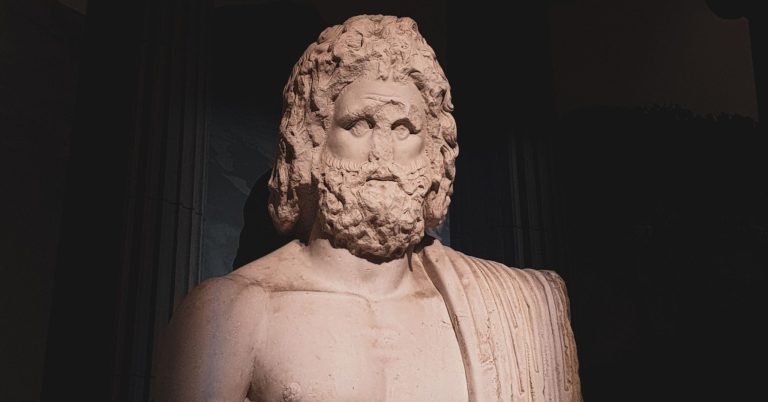 Marble bust of a bearded, muscular male figure draped in a robe over one shoulder, displayed against a dark background.