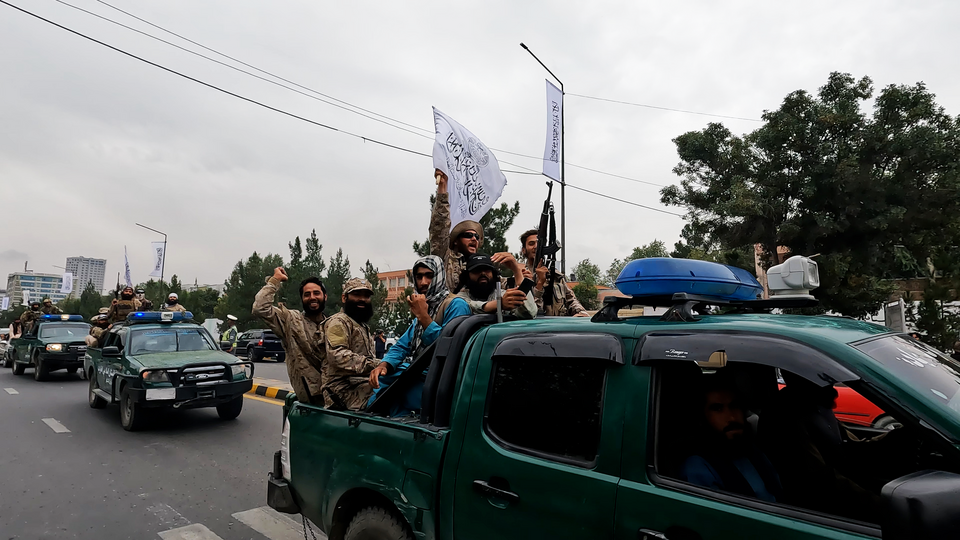 A photograph of green flatbed trucks with blue flashing lights attached parading down the street. In the truck beds are Taliban members - men in combat fatigues, some with flags and guns, some with hats, headscarves or sunglasses. THe mood is celebratory, with one or two in the closest vehicle smiling for the camera. Photograph: Callum Darragh. Source: Flikr via Wikipedia (public domain).