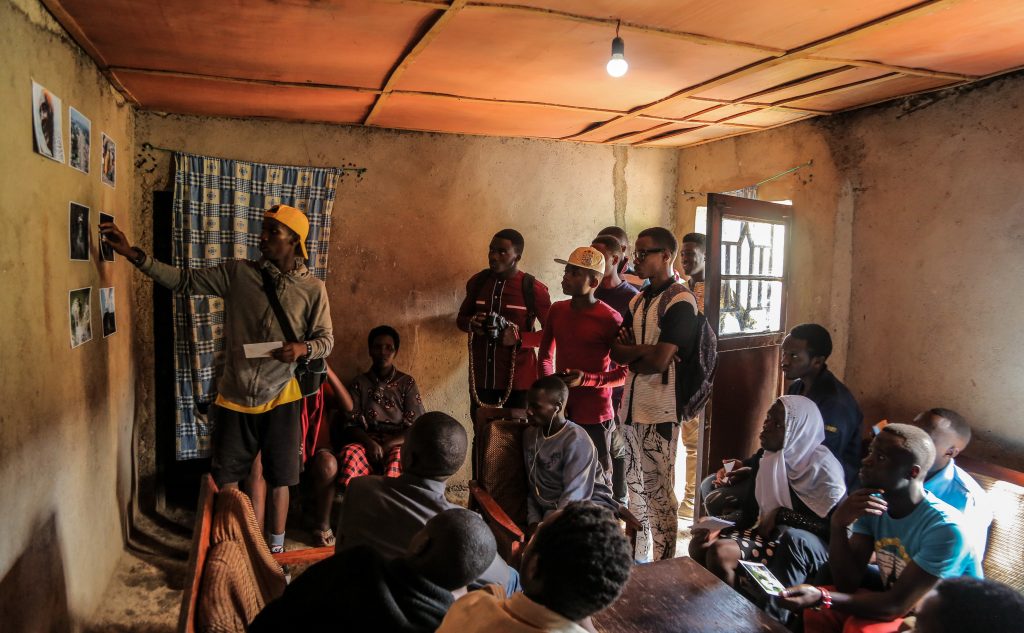 The image shows a group of young people gathered in a modest, dimly lit room with earthen walls and a low ceiling lined with orange panels. A single exposed bulb hangs overhead, casting a warm glow. On the left, a young man in a yellow cap and grey hoodie points to a small grid of taped photographs on the wall. Holding a paper, he appears to lead a discussion or presentation. Around 15 participants—mostly young men, with a few women—watch attentively. Some hold cameras or printed photos, suggesting a photography workshop. Natural light enters through an open doorway on the right.