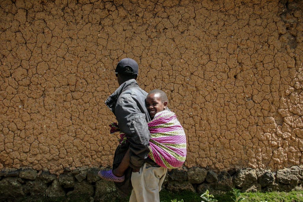 A Black man walks past a sunlit wall of cracked, reddish-brown earth, his back and head facing the camera. He wears a dark cap, oversized grey jacket, and cream trousers. Tied to his back with a vibrant pink, green, and white cloth is a smiling Black child, facing the camera with a joyful grin. The child’s legs stick out from the cloth, wearing purple perforated sandals. The man's stride is casual yet purposeful, while the child’s radiant expression becomes the focal point. The weathered clay wall and scattered greenery contrast beautifully with the colorful fabric and warmth of the child’s smile.
