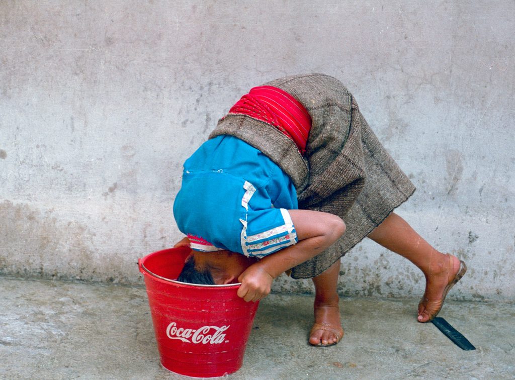 The image features a young girl, of Indigenous descent, bending over a bright red Coca-Cola bucket with her head submerged, arms gripping the rim. She wears a vibrant turquoise blouse with white and blue trim, a brownish-gray skirt, and a red woven sash—suggesting traditional attire. She's barefoot, one foot forward, against a rough light gray concrete wall. The ground is similar but smoother. Natural daylight illuminates the scene. The muted grays and browns of the wall and skirt contrast sharply with the saturated red of the bucket and sash, and the vivid turquoise blouse. The medium shot centers the girl in-frame.