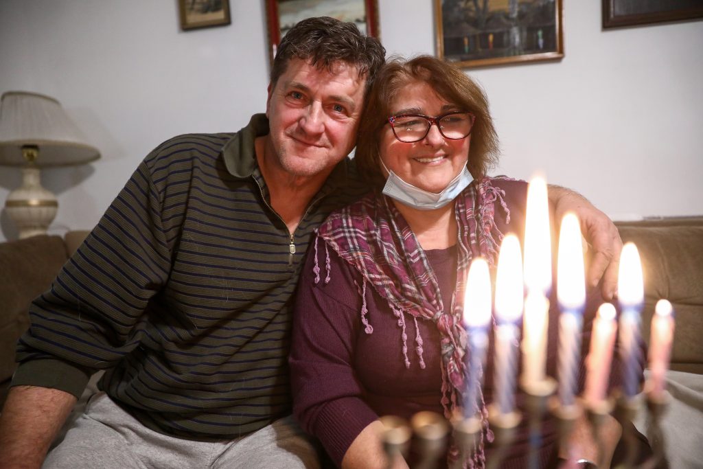 The photograph centers on a middle-aged couple seated closely on a beige sofa. The fair-skinned man, with short dark hair, wears a dark olive striped shirt and gray sweatpants. His arm is draped around his fair-skinned wife, who has shoulder-length light brown hair, dark-framed glasses, a burgundy sweater, and a plaid scarf. A light blue mask rests below her nose as she smiles softly. Between them, a lit menorah glows warmly, with eight visible candles. The home interior features a beige wall with framed art and a cream-shaded lamp. The muted palette includes earth tones and burgundy, creating a cozy, intimate atmosphere.