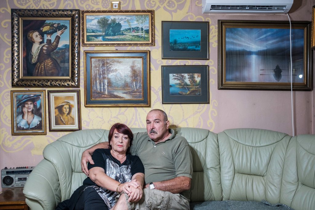 The photograph centers on a middle-aged couple seated on a light sage green leather sectional sofa. The woman, with auburn hair and a pensive expression, wears a dark sleeveless top and skirt, her hands clasped in her lap. Her husband, with short graying hair and a strong build, wears an olive-green t-shirt and khaki shorts, his arm gently around her shoulders. He gazes directly but somberly. Behind them is a pale yellow-cream wall adorned with gold-framed paintings of landscapes, portraits, and still life. A vintage stereo and air conditioner hint at a cozy, lived-in home.