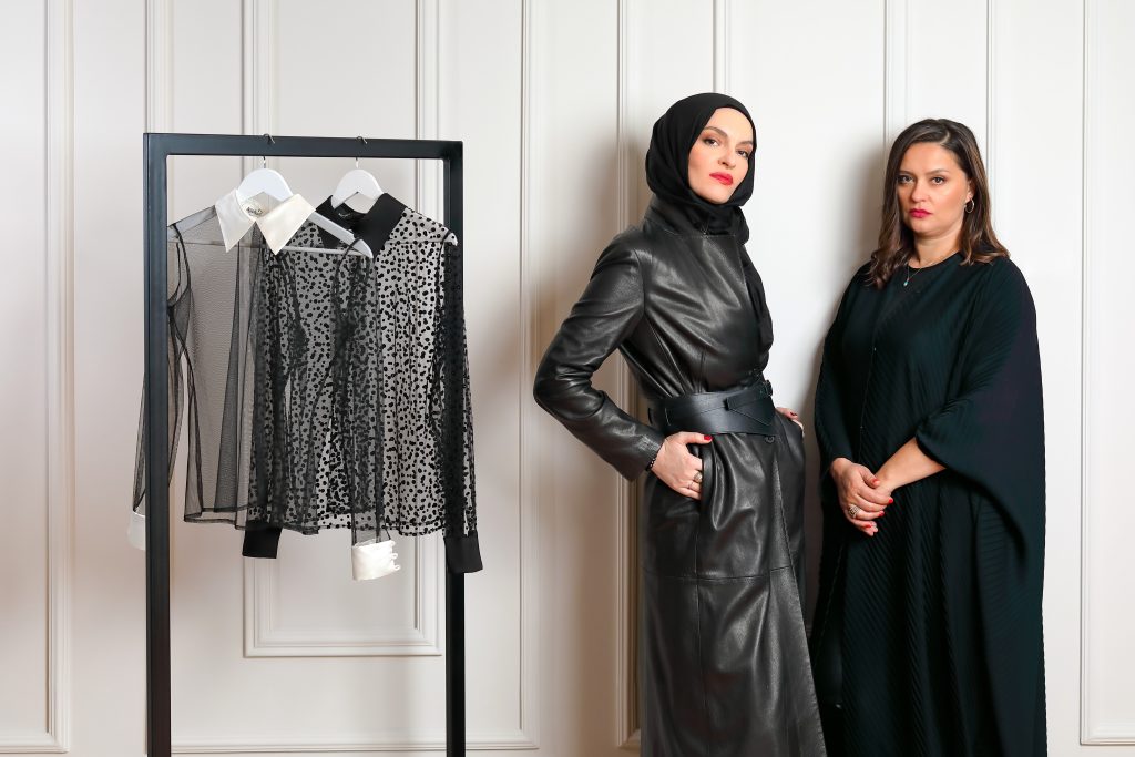 The photograph features two women standing against a white paneled wall. The woman on the left wears a long dark leather coat cinched with a wide black belt, and a black hijab. Her hands are in her pockets, her stance relaxed yet powerful, with a composed, serious expression. The woman on the right wears a dark abaya-style dress with flowing sleeves. She has dark brown hair, wears earrings, and stands with hands clasped, her expression serious but softer. A black clothing rack to the left holds three long-sleeved blouses—white, black, and black with white polka dots. The monochromatic palette conveys elegance and quiet strength.