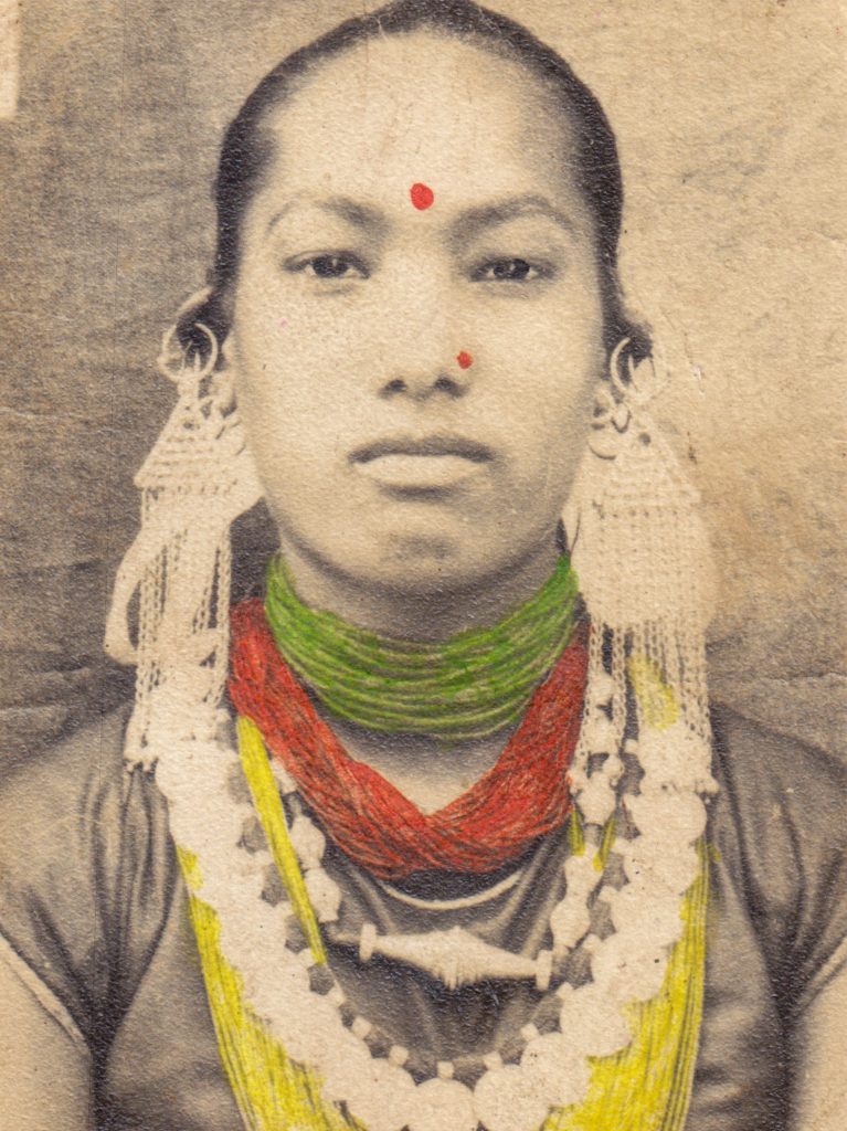 The photograph shows grayscale portrait of a woman in which some parts have been hand coloured. The woman is of apparent Southeast Asian or similar ethnic background. Her dark hair is pulled back and she starts directly into the camera, defiantly. She has a vertical red mark on her forehead, a bindi and is wearing elaborate necklaces and earrings. The necklaces appear to be made of multiple strands of small beads of different colors that have been hand painted on to the photograph in red, green and yellow. The necklaces drape around her neck and chest. Large earrings are visible as well. The image has a vintage aesthetic and feel.