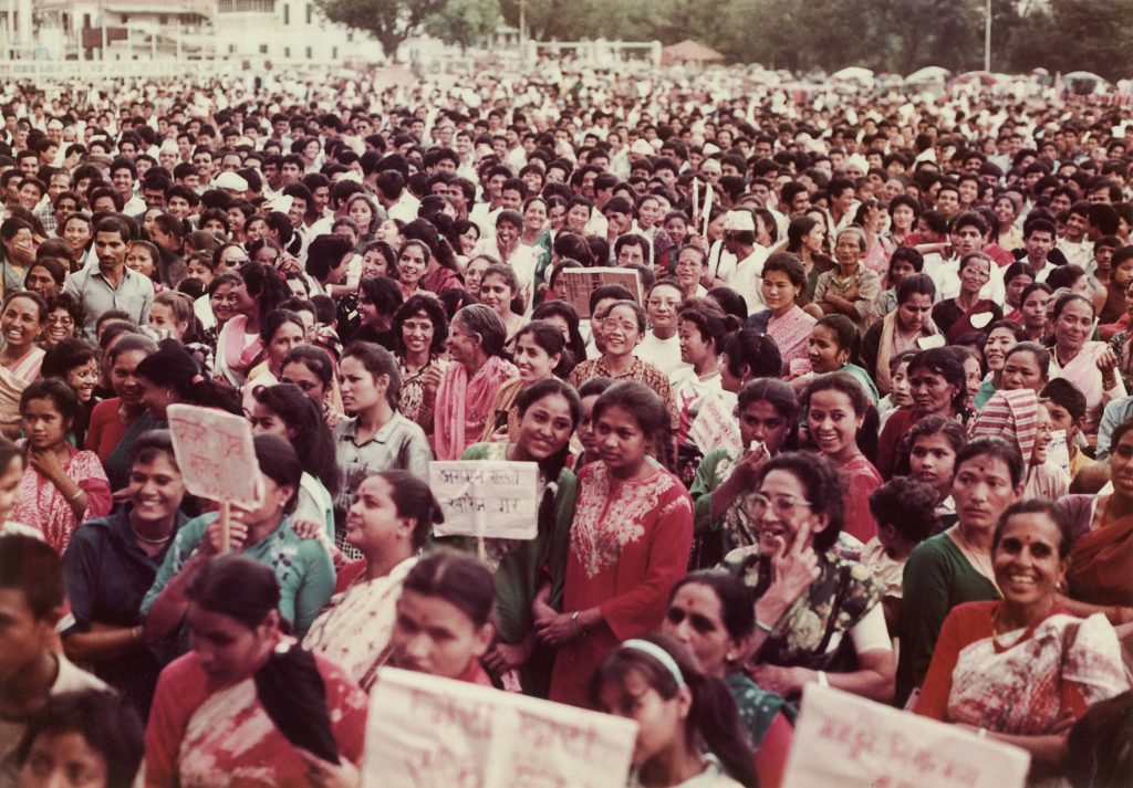 The photo shows a large crowd of standing women stretching far back. They are all facing in the same direction and some hold plackards. Many of the women are smiling and chatting to each other. The scene looks convivial. 