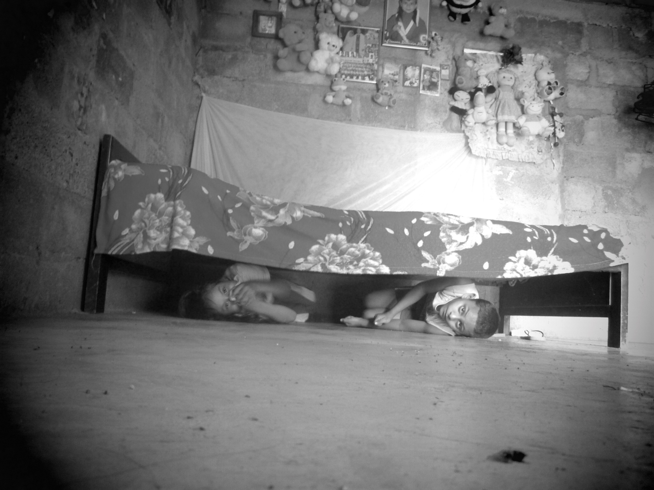 The low-angle black-and-white image shows two children hiding under a bed, viewed from floor level. One, slightly older, peers out while the other covers their face, seemingly suppressing a giggle. A floral bedspread drapes from a simple wooden frame in the corner of a sparse room, its rough, bare brick walls suggesting poverty. Above the bed hang stuffed animals, framed photos, and a decorative wall hanging. The children’s expressions mix playfulness and apprehension. The monochrome palette heightens the nostalgic, slightly melancholic mood, contrasting innocence and hardship in the scene’s intimate, candid moment.